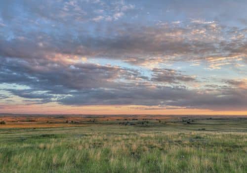 A photo of the Eastern Colorado plains, with green grass, brown, rolling hills in the distance, and clouds in a blue and pink sky