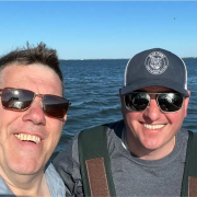 Mike and Ray, owners of the oyster farm, in sunglasses, standing on a boat smiling into the camera.