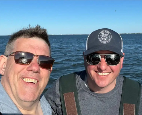 Mike and Ray, owners of the oyster farm, in sunglasses, standing on a boat smiling into the camera.