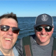 Mike and Ray, owners of the oyster farm, in sunglasses, standing on a boat smiling into the camera.