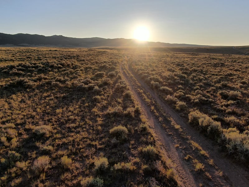 a road leads through rangeland to distant mountains with sun on the horizon