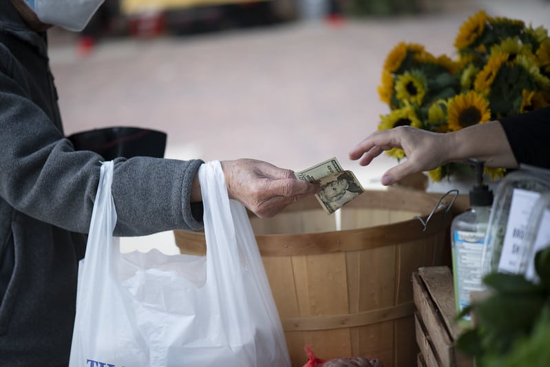 arm holding a bag hands off money to a reaching hand at a farmers market booth