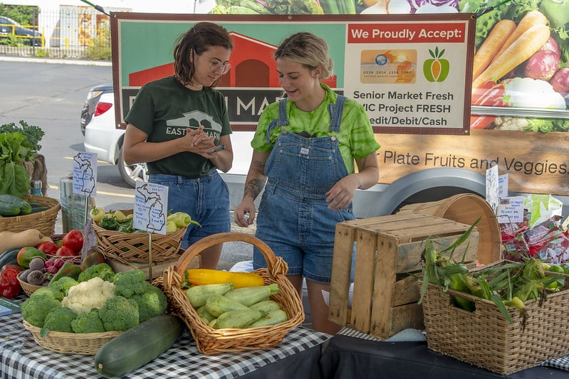Two people at a farmers market stand with a variety of vegetables on display.