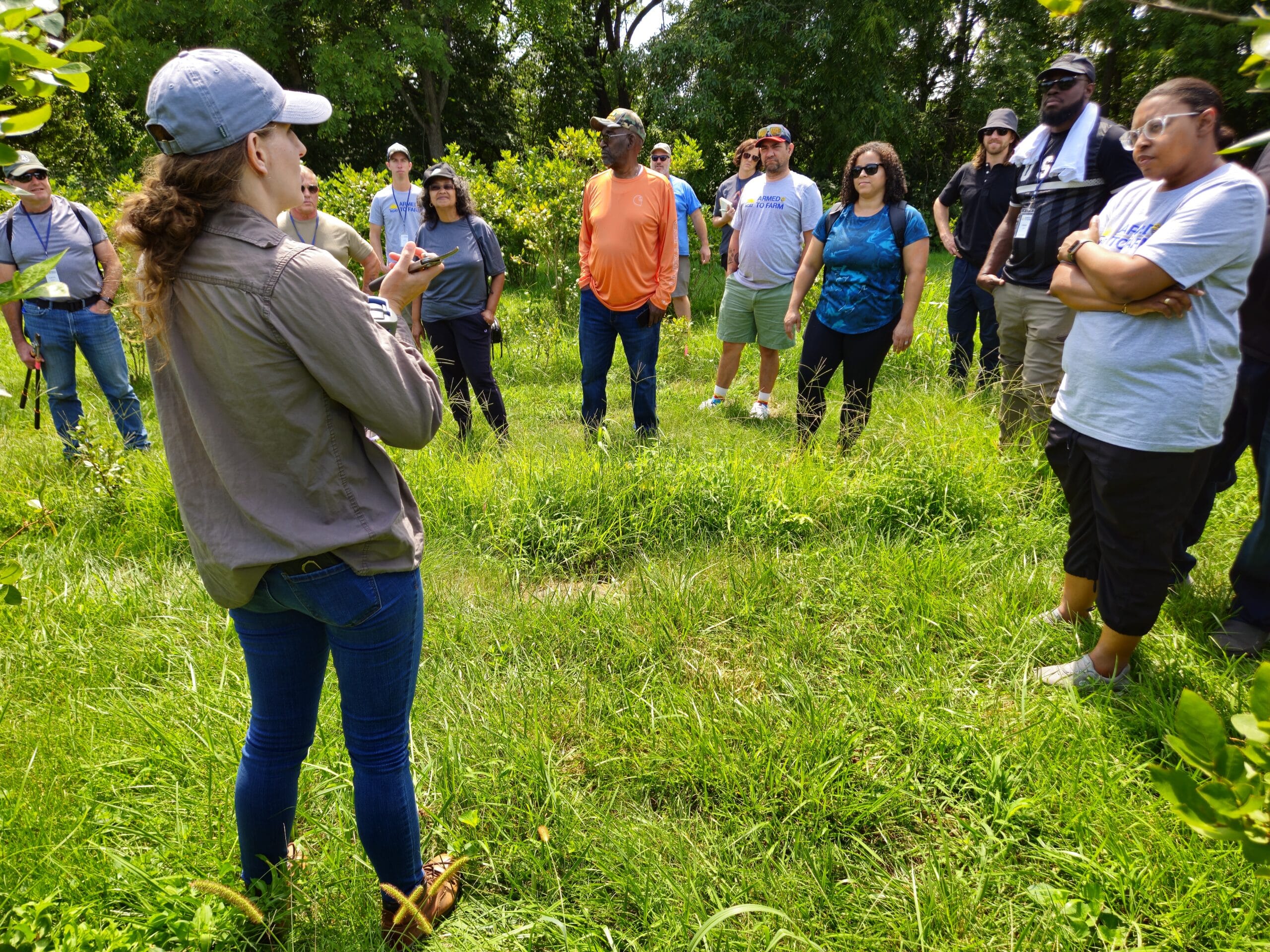 Un grupo de personas del público está parado al aire libre sobre el césped, escuchando a un presentador