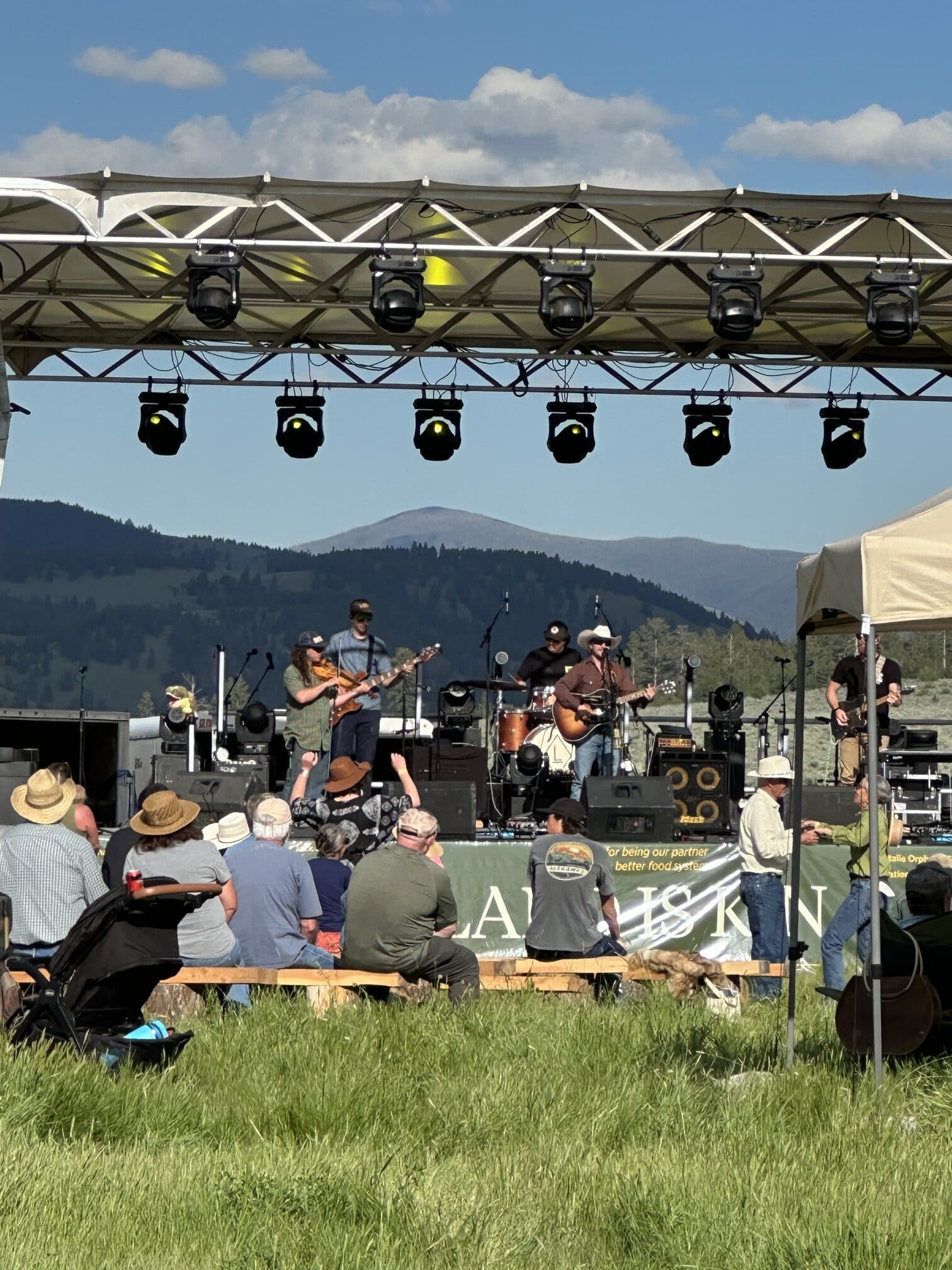 presentación musical en un escenario al aire libre frente a las montañas, con el público sentado en bancos